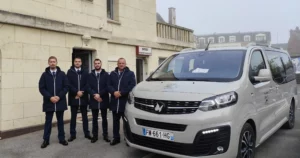 Quatre porteurs funéraires posant devant une chambre funéraire à côté d'un corbillard gris.
