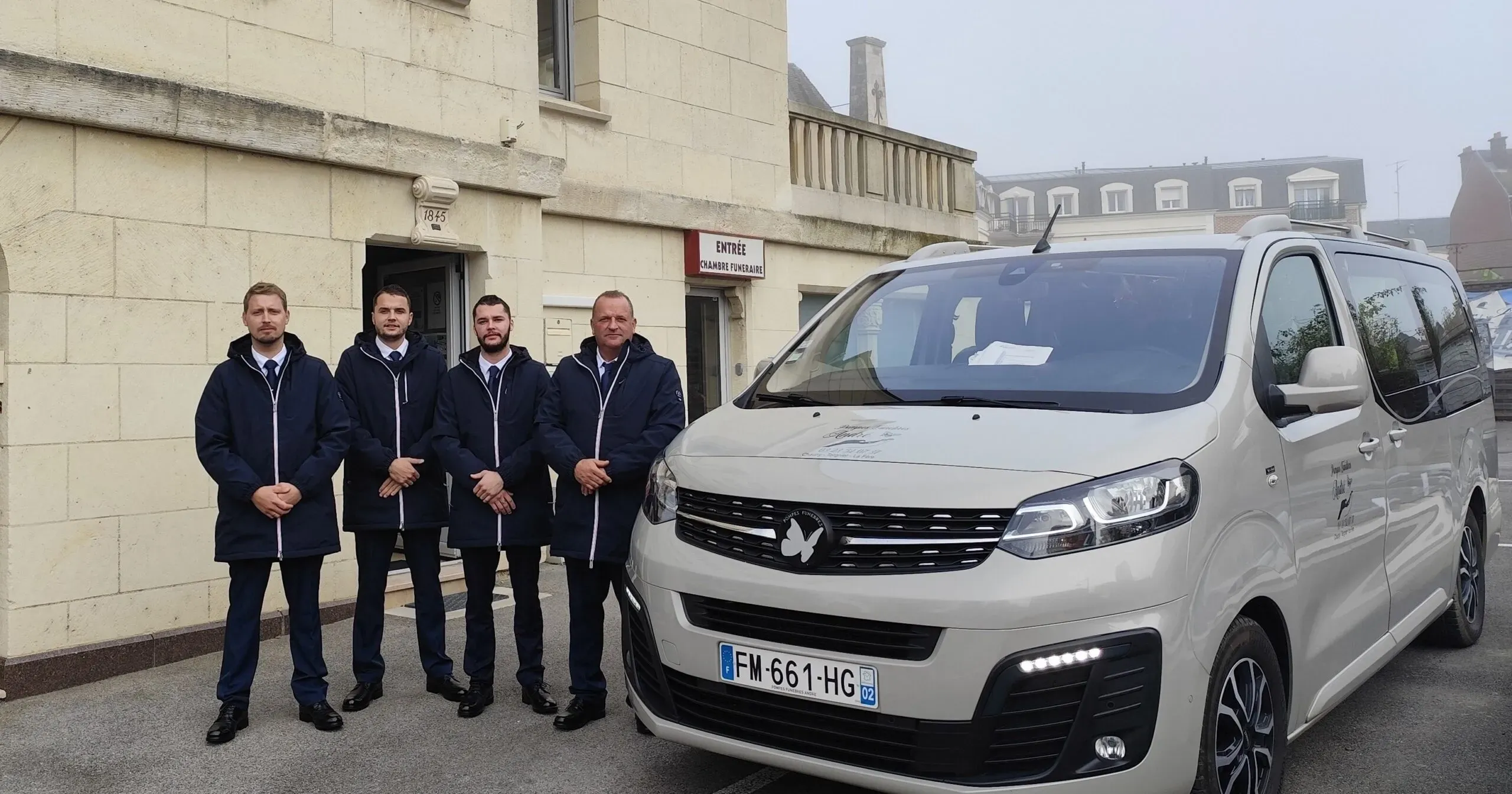 Quatre porteurs funéraires posant devant une chambre funéraire à côté d'un corbillard gris.